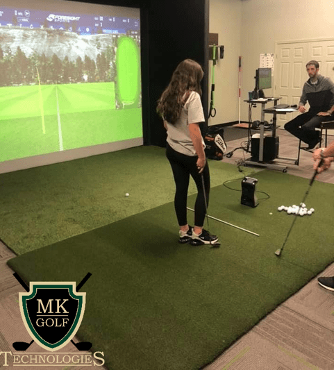 Player standing on an artificial turf mat, preparing to swing at a golf simulator projected screen; instructor and observer nearby in a room with golf equipment visible.