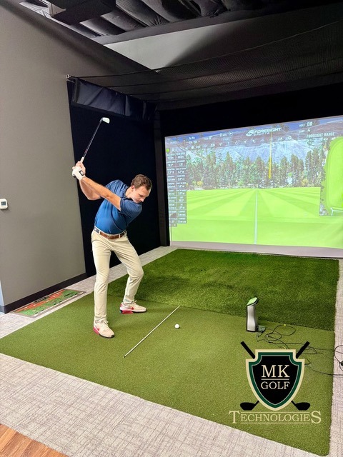 Man in a blue shirt swinging a golf club in an indoor simulator bay, a large virtual course on screen behind him